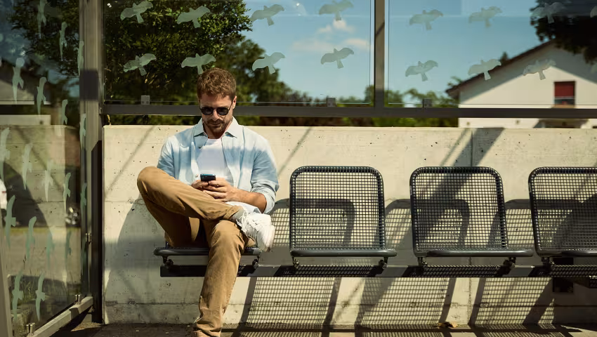 Mann sitzt mit einem Smartphone in der Hand auf einer Bank an einer Bushaltestelle.