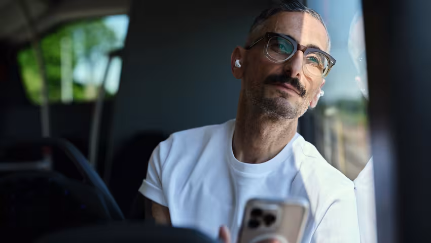 Mann schaut lächelnd aus einem Busfenster und hält dabei ein Smartphone in der Hand.