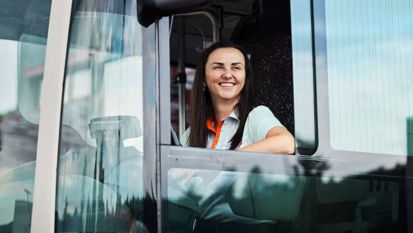 Busfahrerin sitzt am Steuer und blickt lächelnd aus dem geöffneten Fahrerfenster.