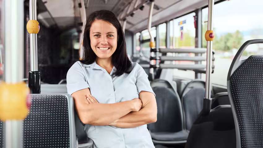 Busfahrerin steht mit verschränkten Armen und freundlichem Lächeln im Innenraum eines Busses.