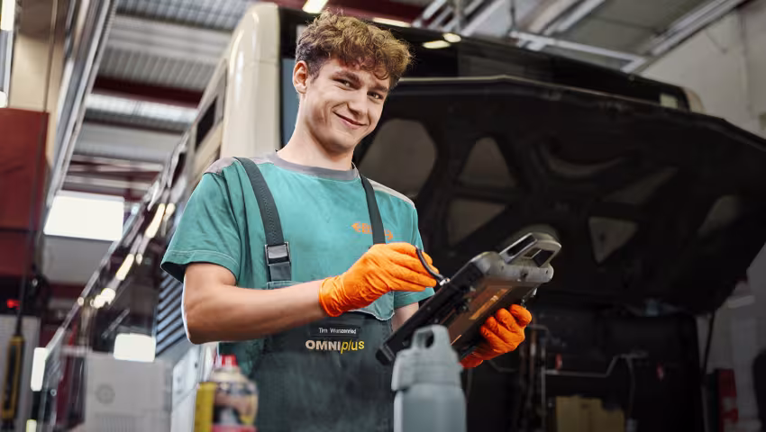 Ein Lernender Automobil-Mechatroniker steht lächelnd in einer Werkstatt.